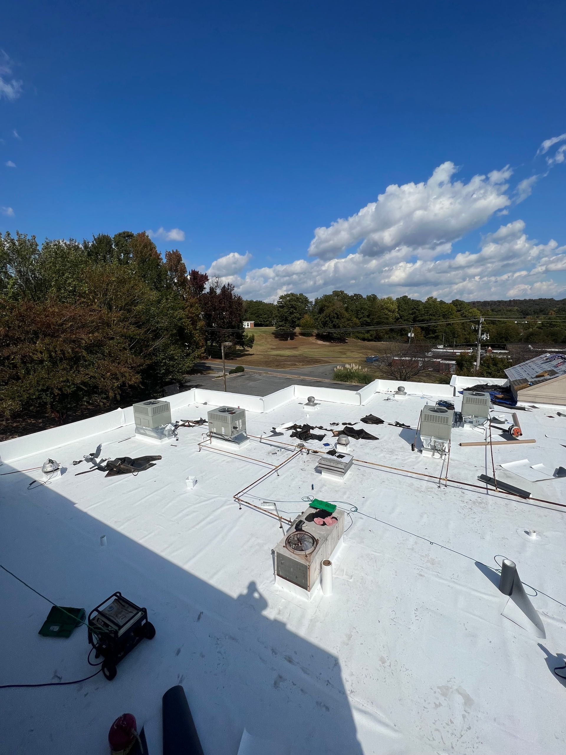 A high-angle view of a flat, white roof with HVAC units under a bright blue sky with scattered clouds.