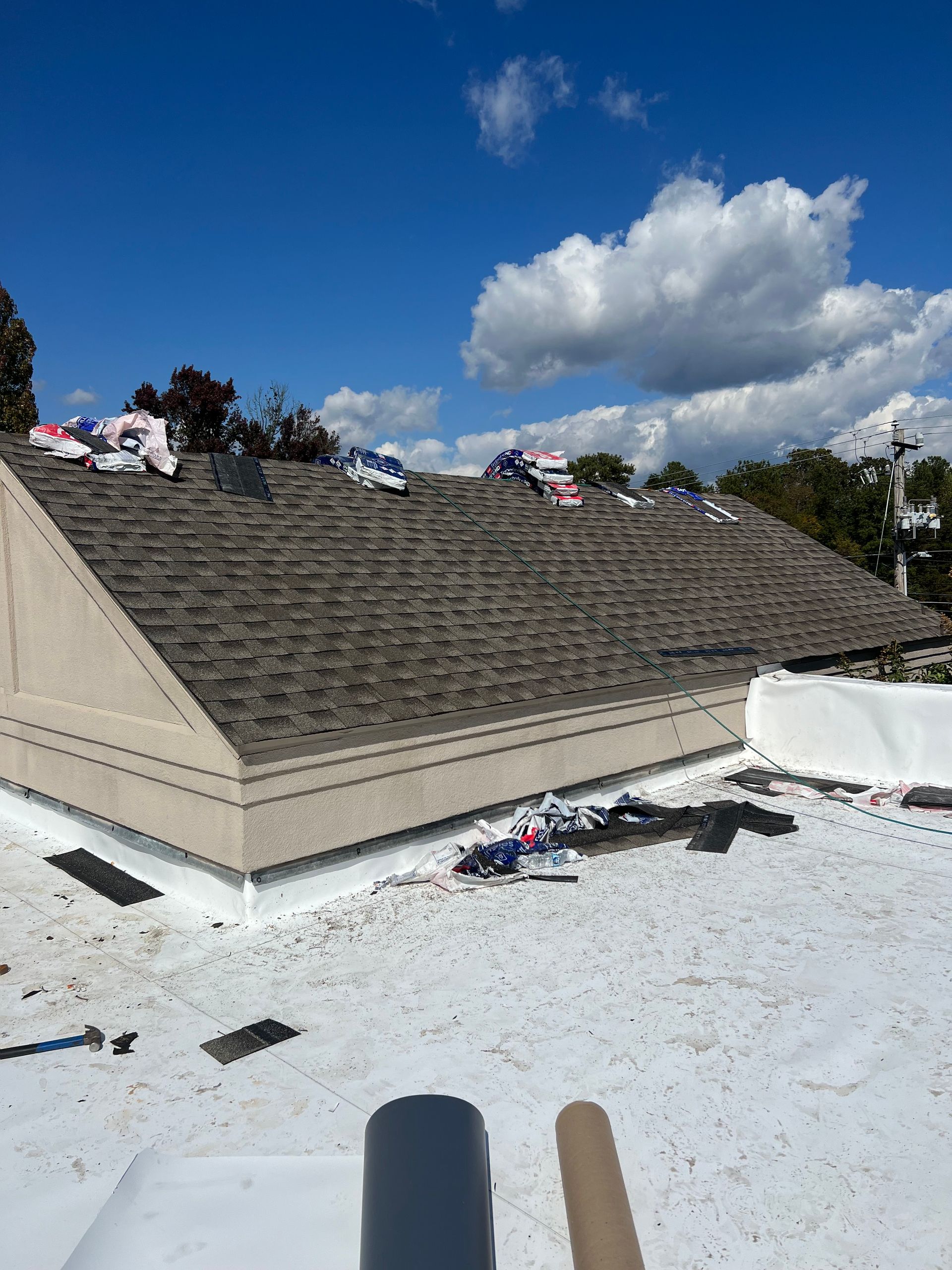 A low-angle view of a building roof under construction, showing shingles on a slope above a flat white roofing membrane.