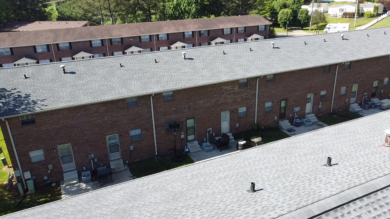 An aerial view of a long, multi-unit brick apartment building with a grey shingled roof and rows of individual entrances.