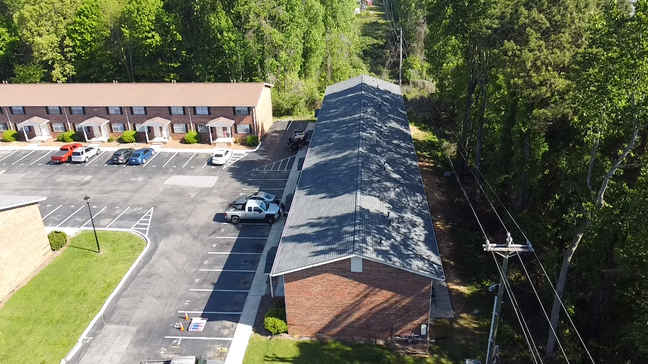 An aerial view of an apartment complex, showing two brick buildings, a parking lot with several cars, and nearby trees.