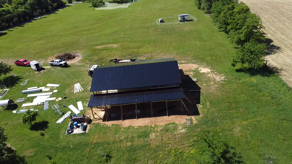 Aerial view of a barn with a newly installed dark solar roof panel array, surrounded by a grassy field and construction.