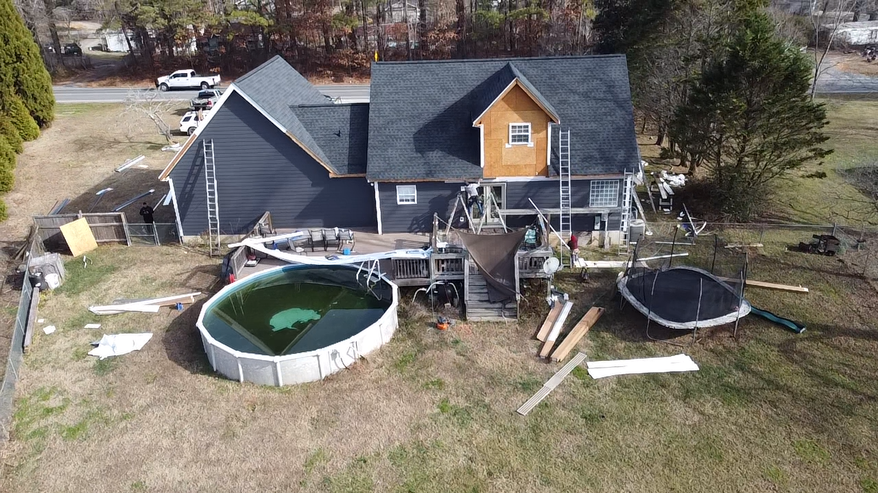 Aerial view of a house undergoing exterior renovations, with a large, green-water pool and a trampoline in the backyard.