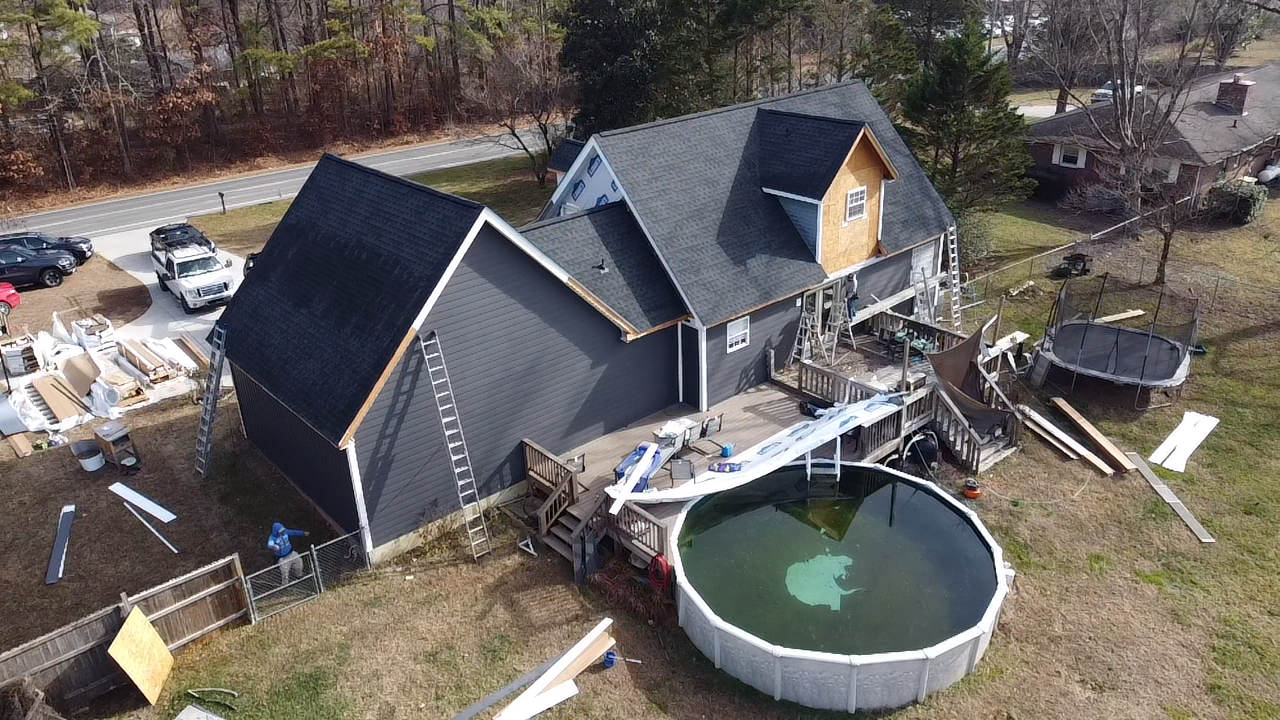 An aerial view of a dark-sided house under renovation with a deck, a round above-ground pool, and construction materials.