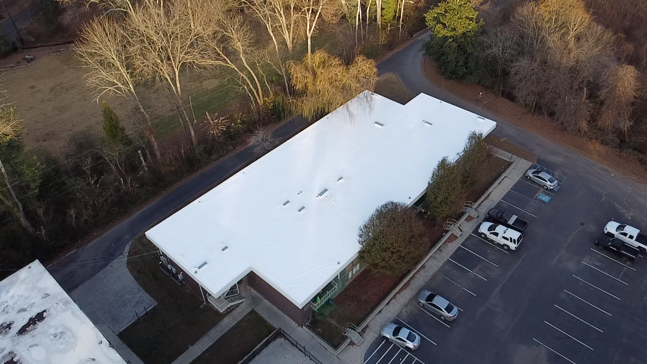 An aerial view of a building with a large white flat roof, surrounded by trees and a parking lot with several cars.