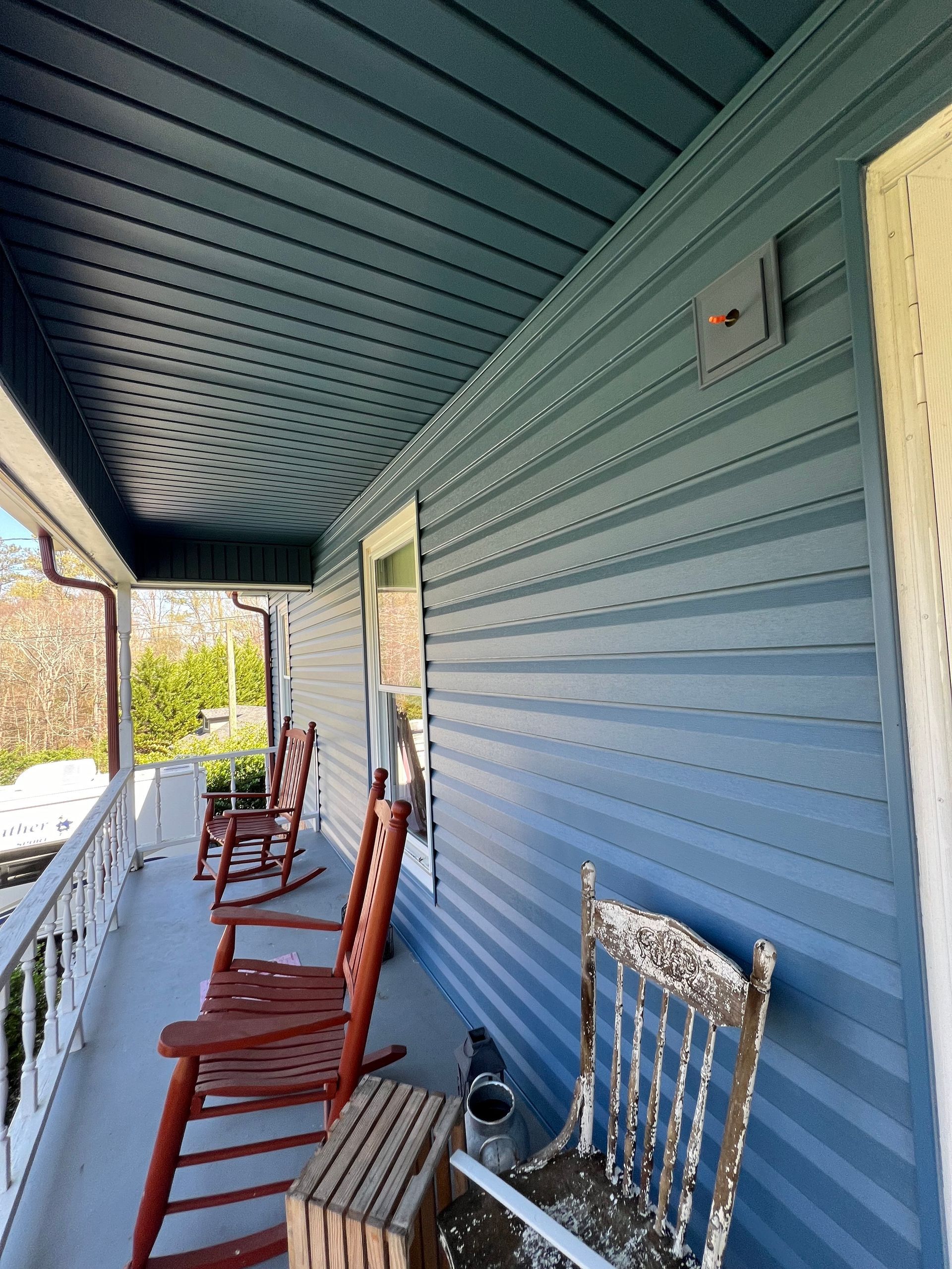 A covered porch featuring blue vinyl siding, wooden rocking chairs, and a weathered chair on a concrete floor.