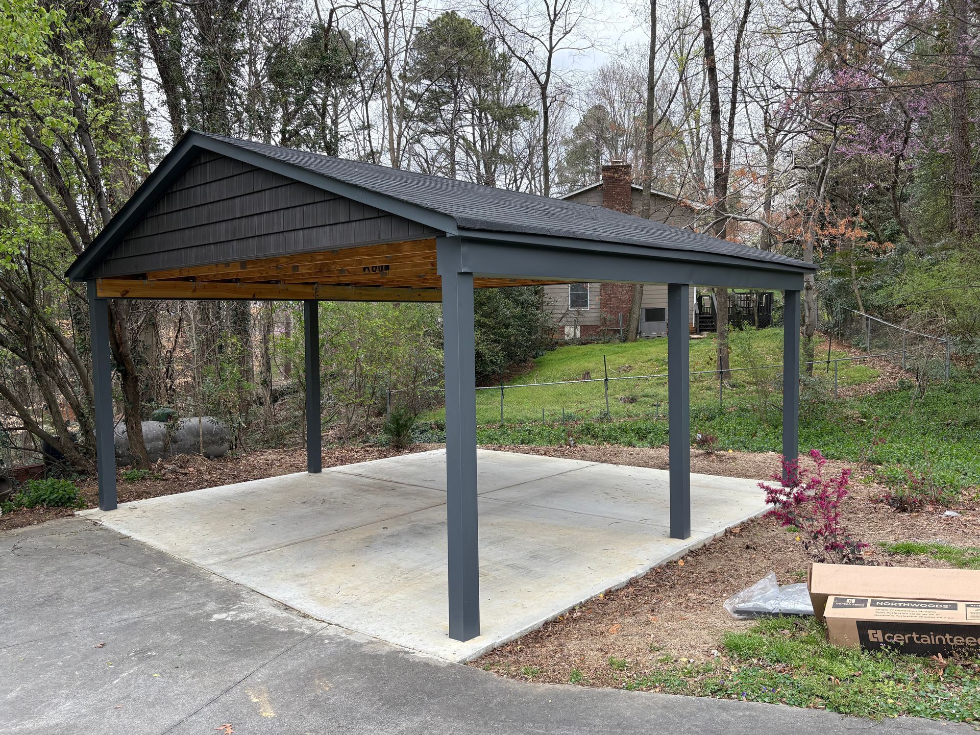 A dark grey wooden carport structure stands over a concrete pad in a yard with trees in the background.