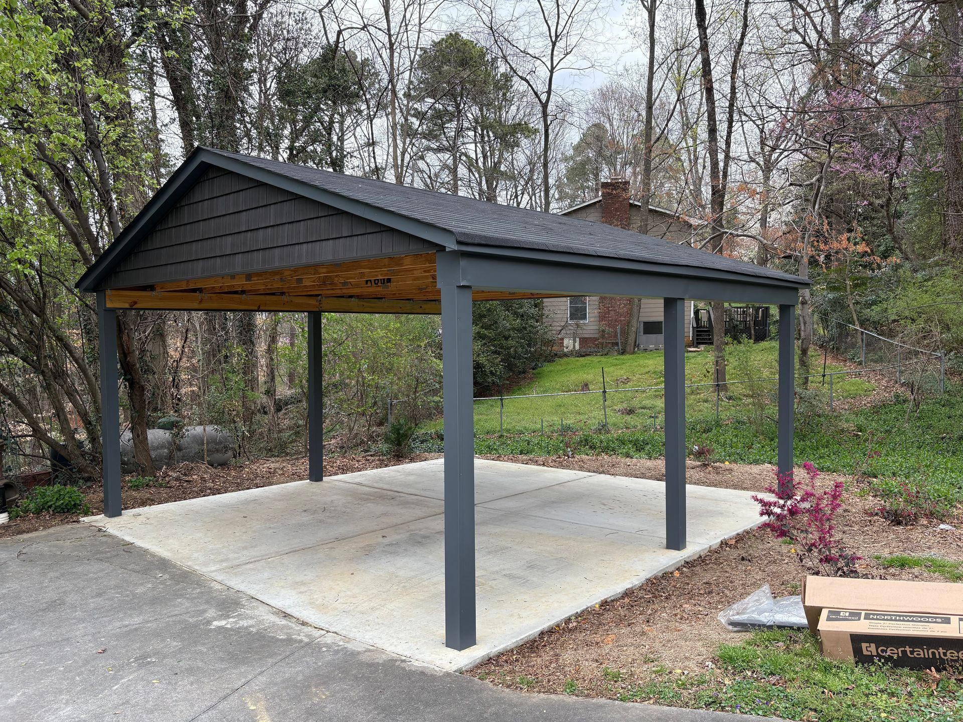 A dark gray, open-sided metal carport with a shingled gable roof stands on a concrete slab in a wooded yard.