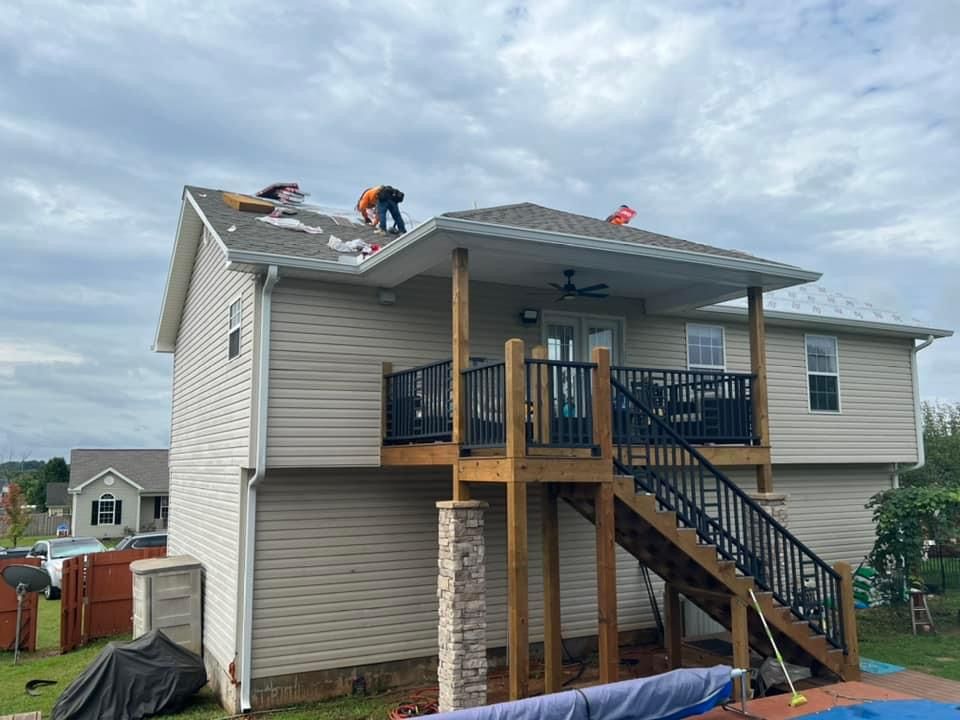 Two workers repair the roof of a two-story beige house with a raised wooden deck and stairs in the backyard.