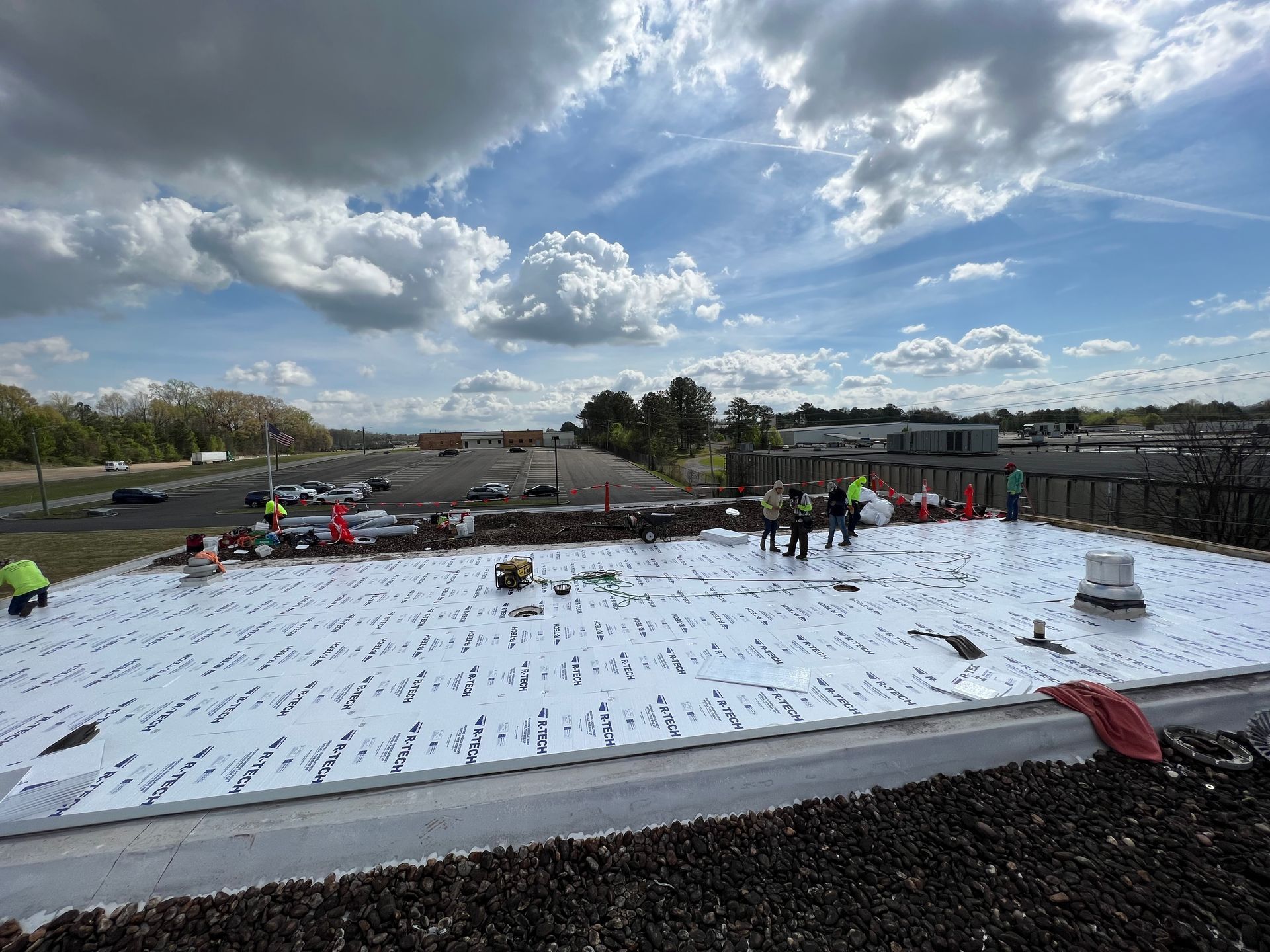 Construction workers install white insulation panels on a flat roof under a blue sky with scattered clouds.