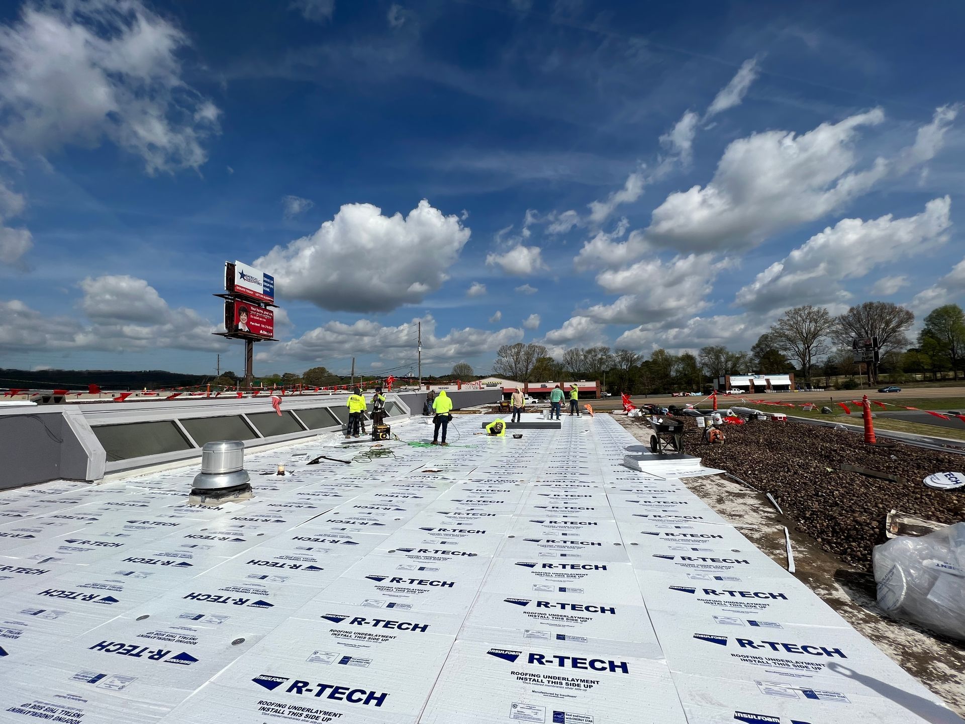 Workers in high-visibility vests install R-Tech insulation boards on a commercial flat roof under a blue, cloudy sky.