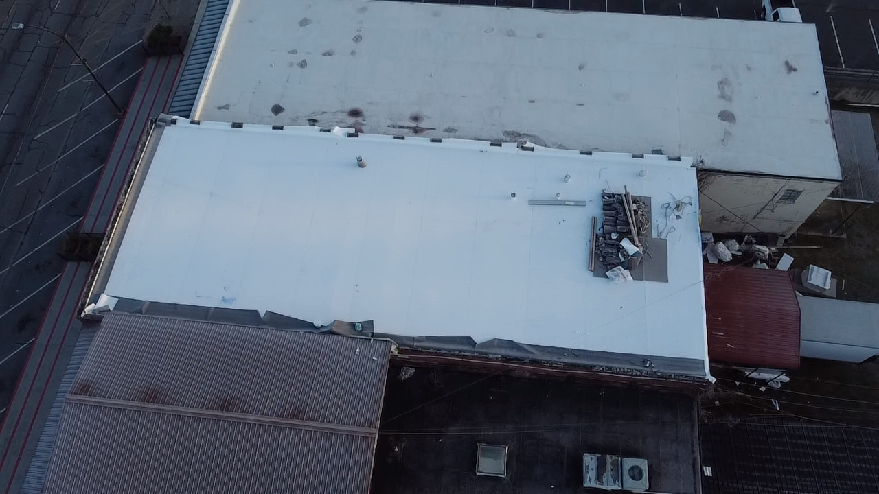 An aerial view of a building with a large, white flat roof, adjacent parking lots, and smaller roof sections.