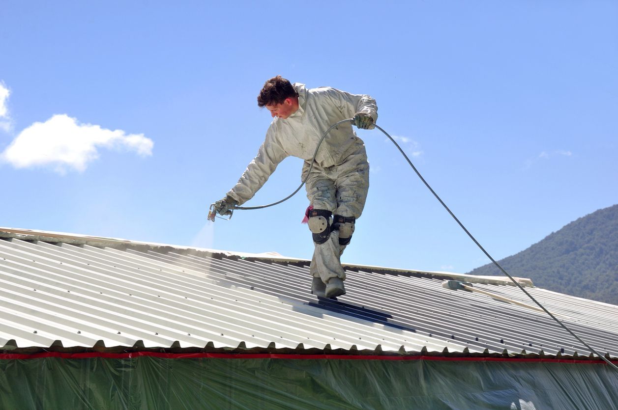 A person in protective coveralls uses a spray gun to apply a white coating to a corrugated metal roof against a blue sky.