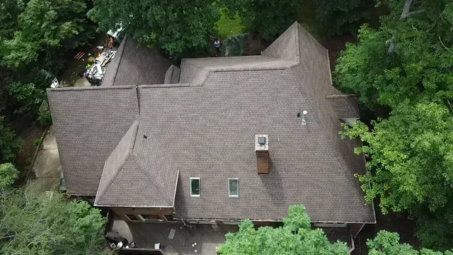 A high-angle aerial view of a brown shingled roof with a chimney and two small windows, surrounded by lush green trees.
