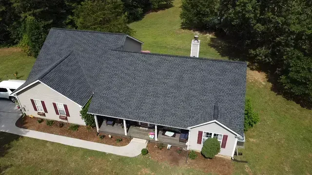 An aerial view of a single-story house with a grey shingled roof, red shutters, and a front porch, set on a grassy lot.