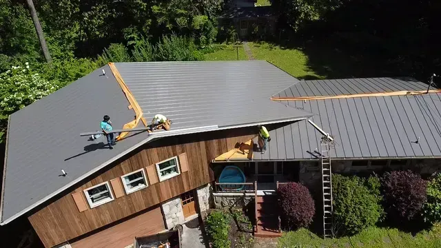 Construction workers installing a gray metal roof on a house with wood siding and stone accents on a sunny day.