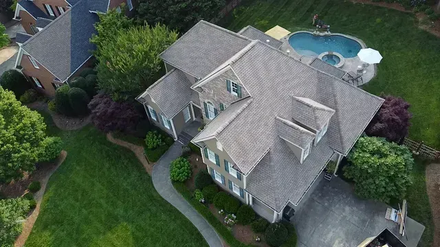 An aerial view of a two-story suburban house with a grey roof, front walkway, pool in the backyard, and surrounding trees.