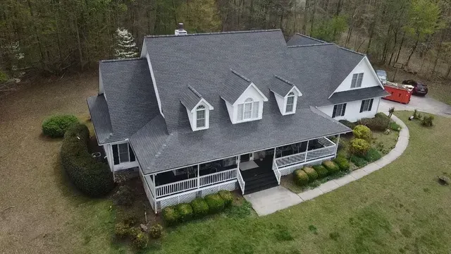 An aerial view of a white, two-story house with a grey roof, front porch, and dormer windows, surrounded by trees.