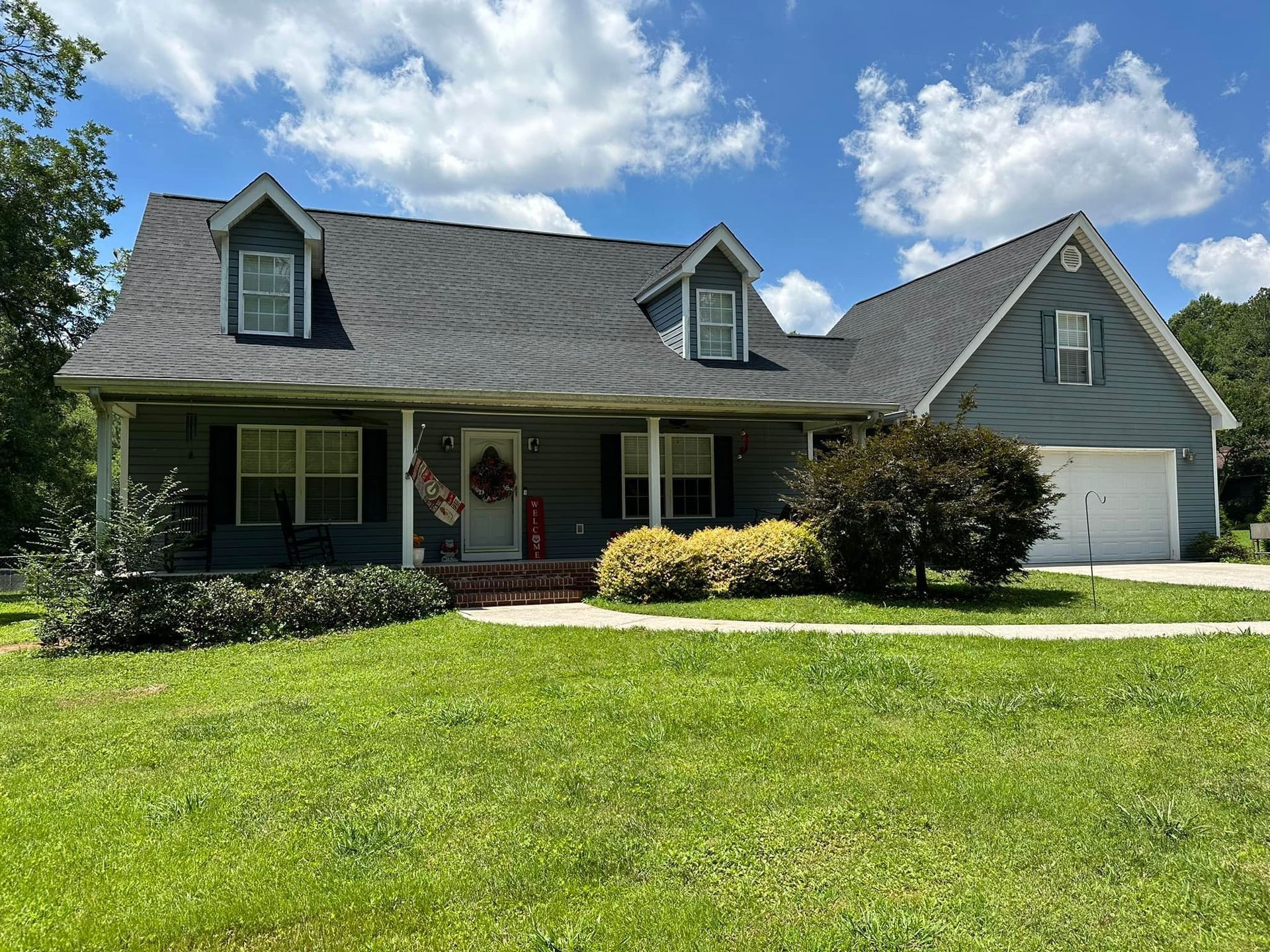 A blue two-story house with a white garage, dark roof, front porch, and a grassy front yard under a blue sky.