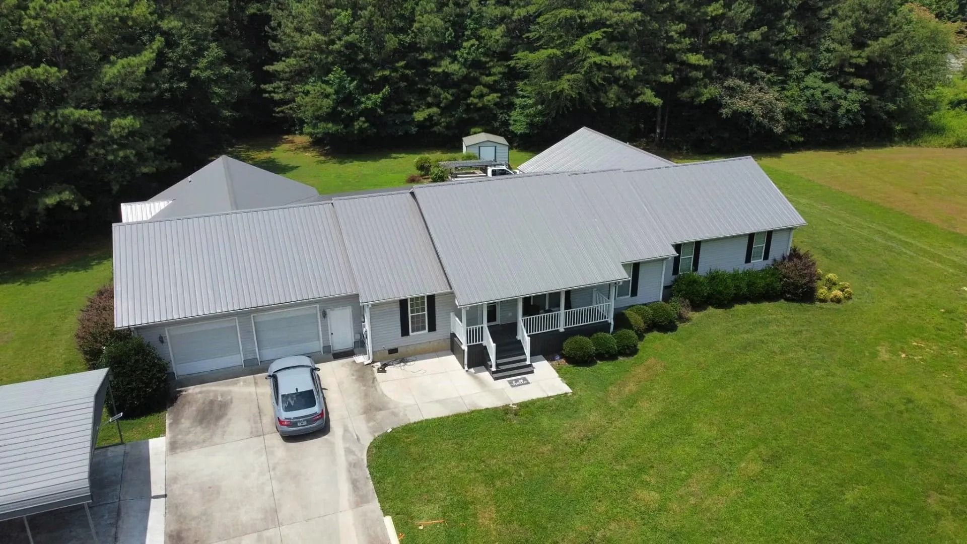 An aerial view of a light-gray ranch-style house with a metal roof, a paved driveway, and surrounding green lawn and trees.