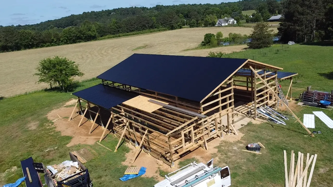An aerial view of a wooden post-frame barn under construction in a grassy field, featuring partially installed black roofing.