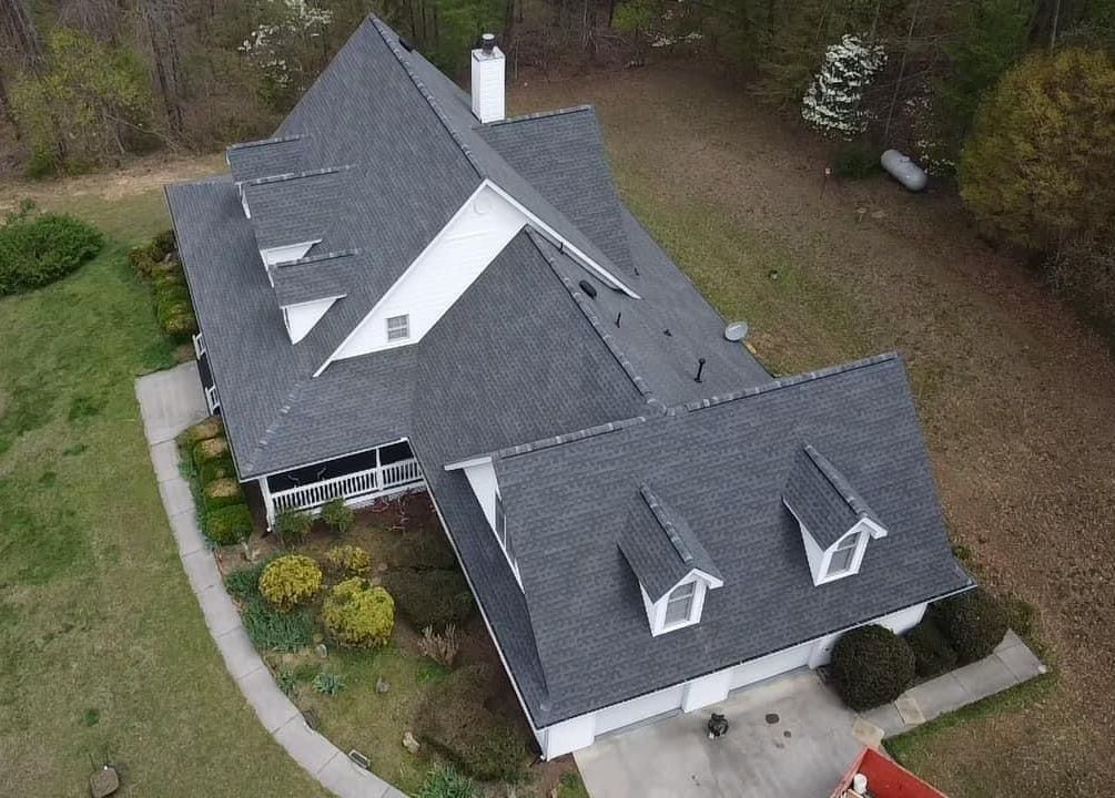 An aerial view of a house with a dark gray shingled roof, white accents, and two dormer windows over a concrete driveway.