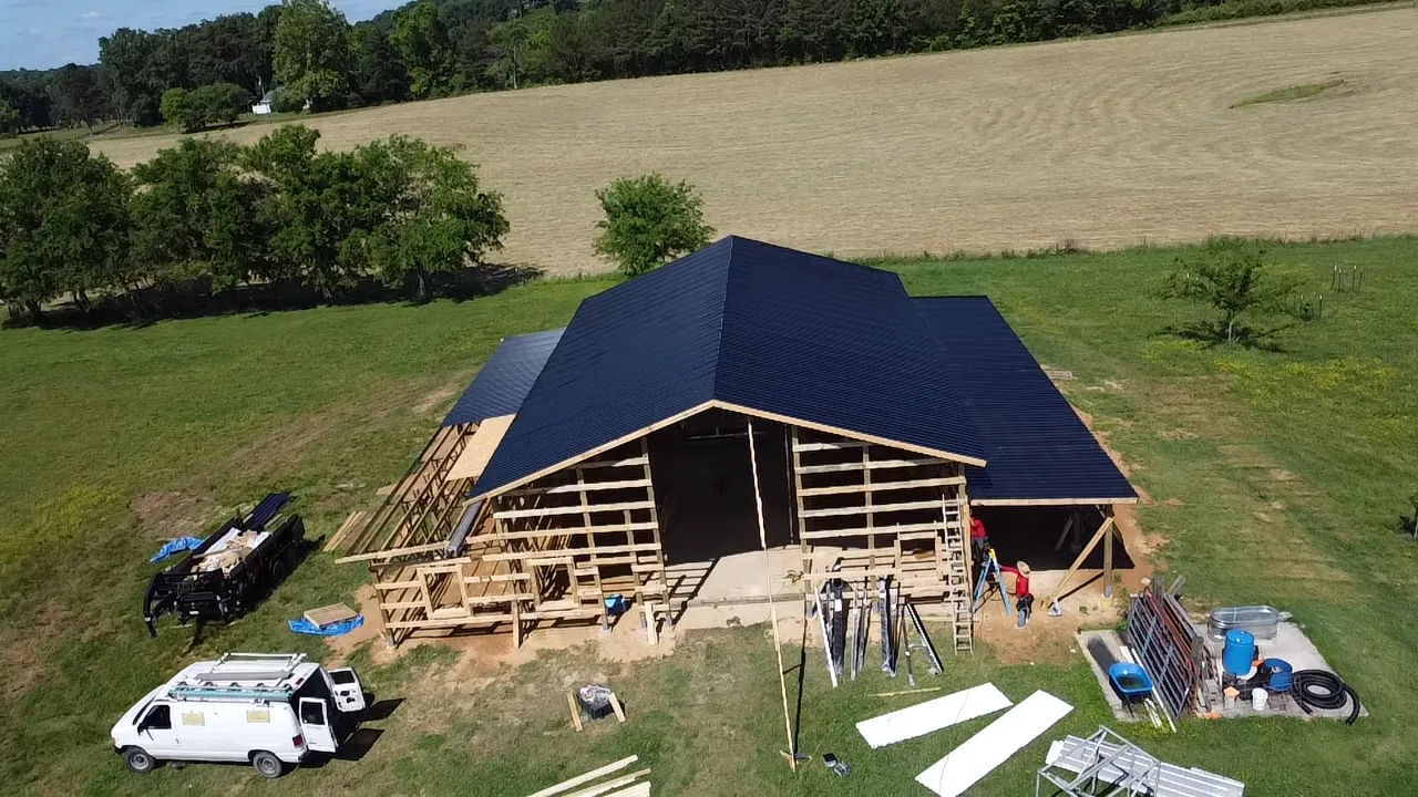 An aerial view shows workers renovating a wooden barn with a newly installed black roof on a sunny, rural property.