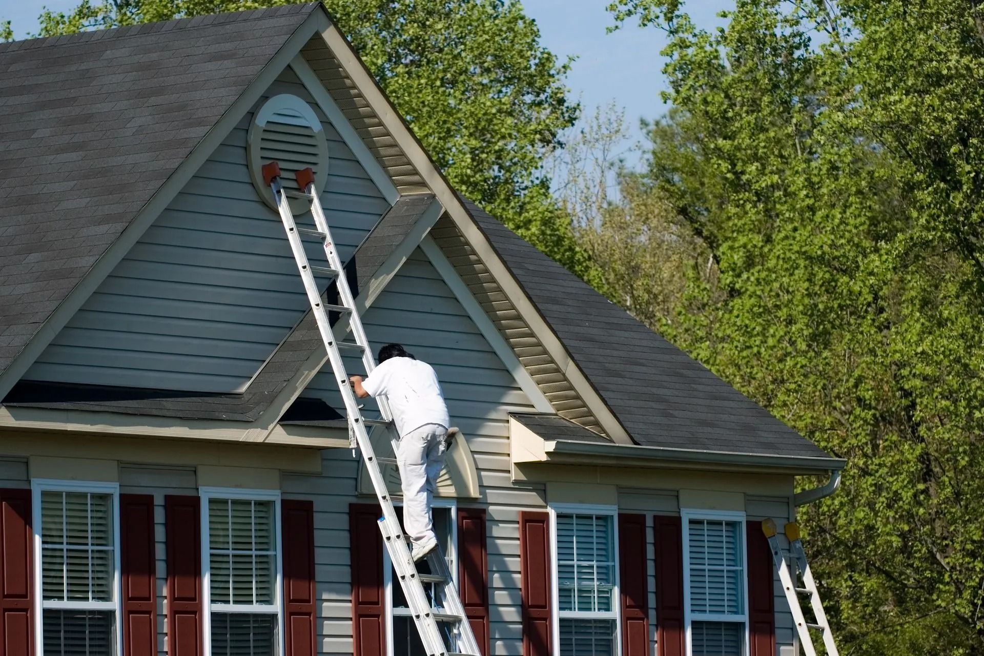 A person in white clothing climbs a ladder leaned against the side of a house to reach an attic vent.