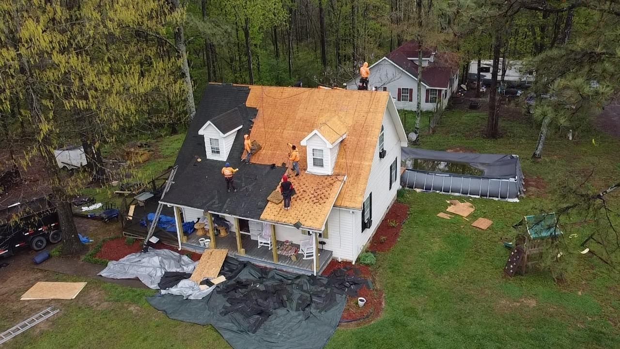 An elevated view of workers replacing a roof on a white house, with new dark shingles on one side and bare wood on the other.