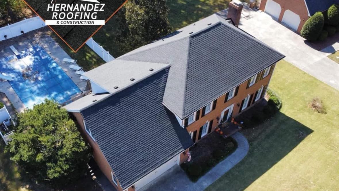 An aerial view of a brick house with a newly installed dark shingled roof next to a backyard swimming pool.