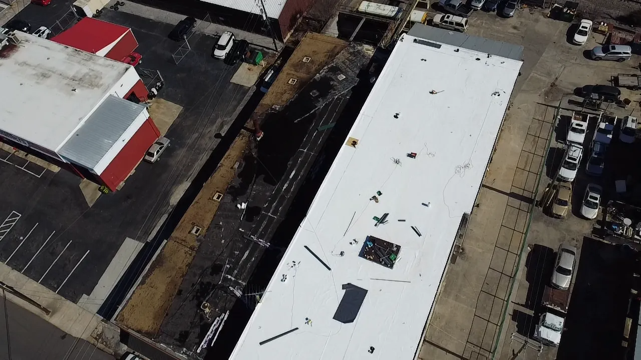 Aerial view of a white commercial building roof under construction alongside a parking lot and a brown dirt excavation.