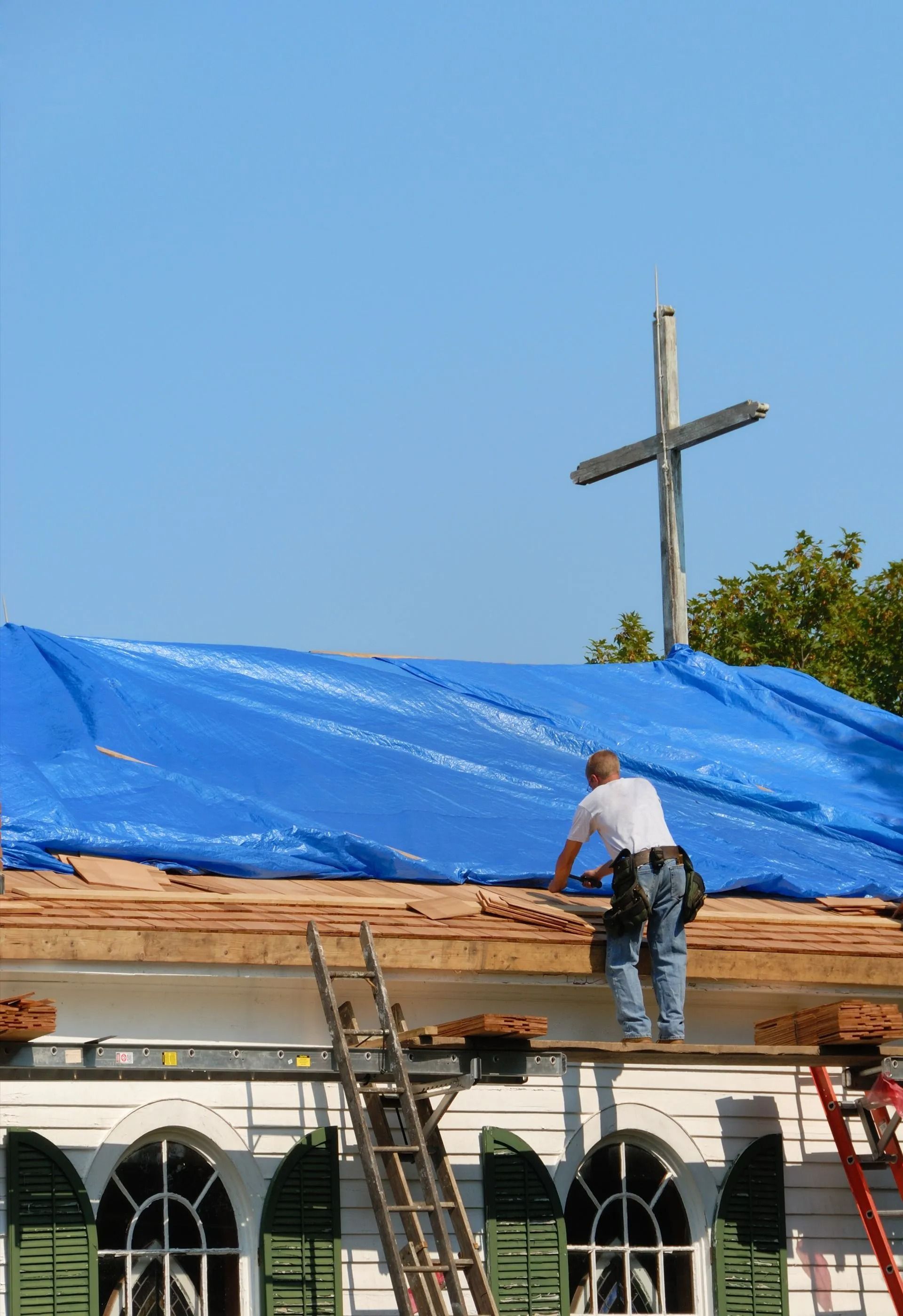 A worker on scaffolding repairs a roof covered with a blue tarp, beneath a large wooden cross on a sunny day.