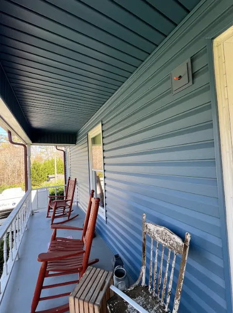 A side view of a porch with blue siding, two wooden rocking chairs, a white weathered chair, and a view of trees outside.