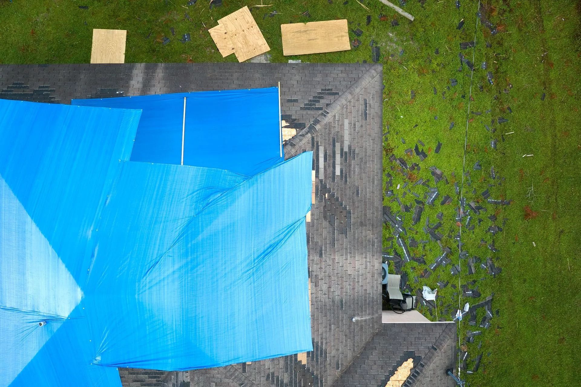 An aerial view of a house roof partially covered with blue tarps, with debris scattered on the lawn below.