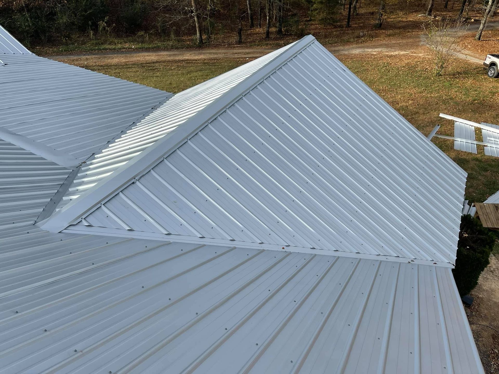 A high-angle view of a grey metal roof with a valley and hip section, set against a background of trees and a field.