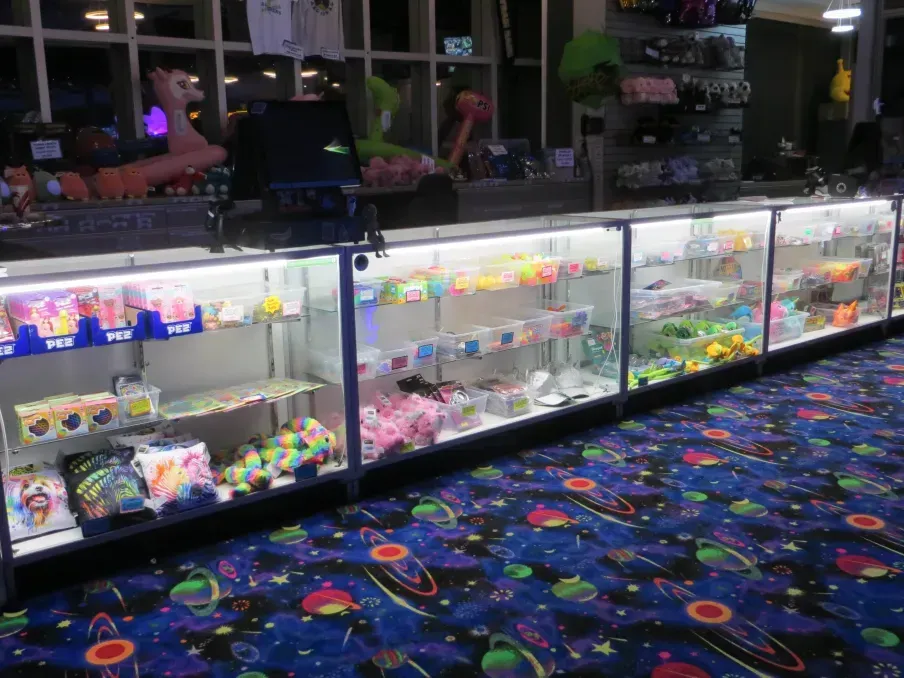 An arcade prize counter with glass display cases filled with toys and candy, set on a space-themed carpet.