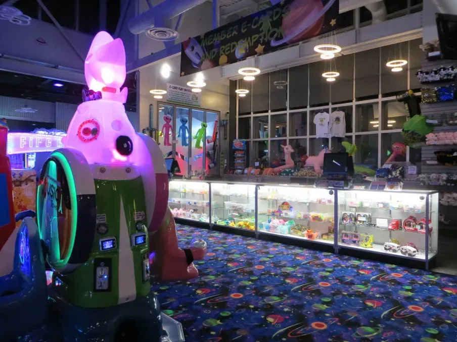 An arcade with colorful games, prizes, and a prize counter under bright lights; a robot game is in the foreground.
