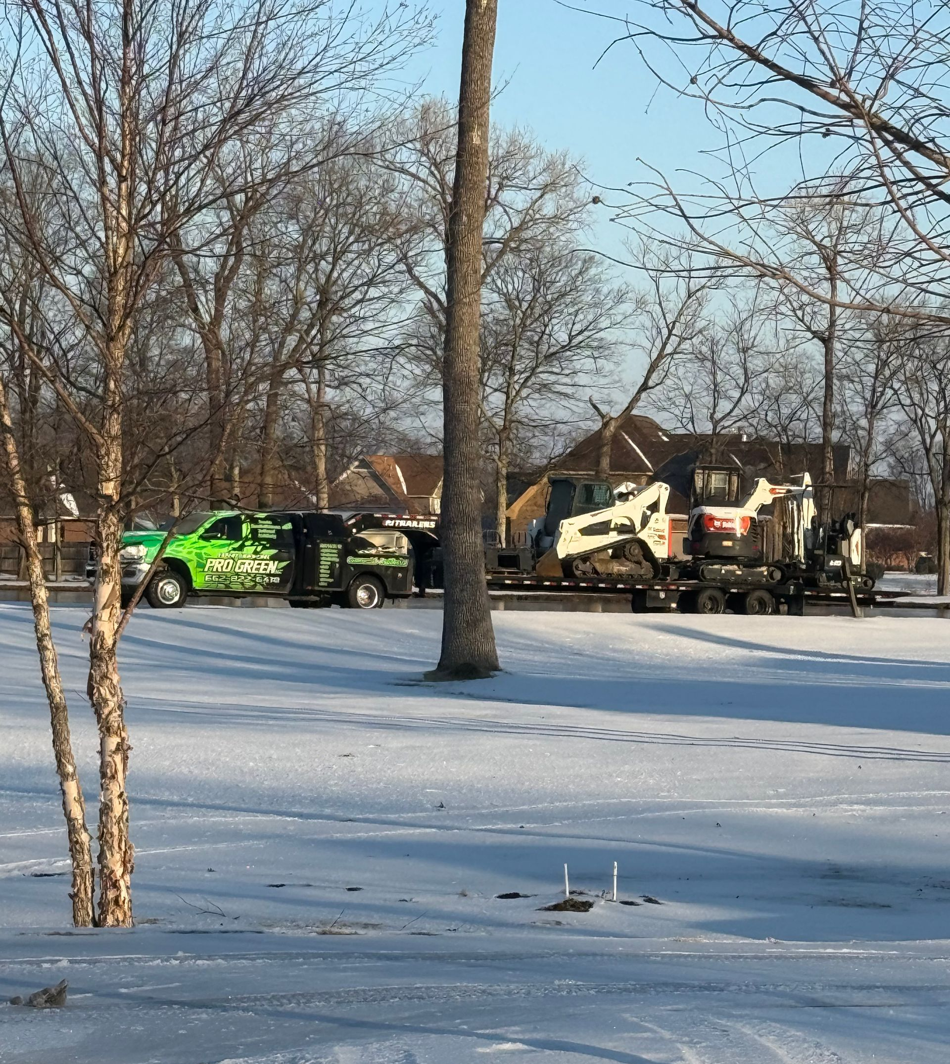 Snowy scene with green and black work trucks, a Bobcat on a trailer, and trees.