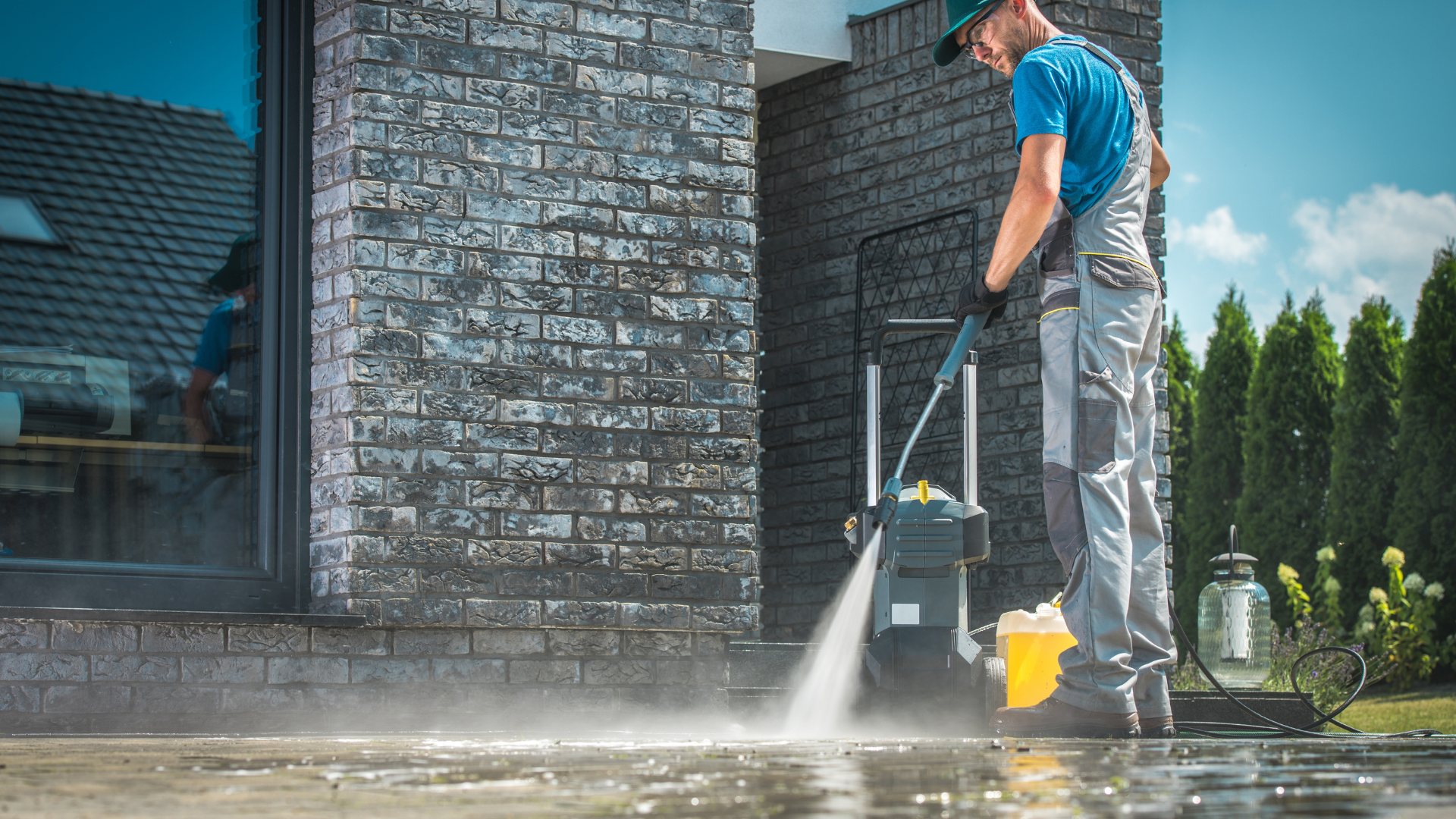 A man is using a high pressure washer to clean the sidewalk in front of a brick building.