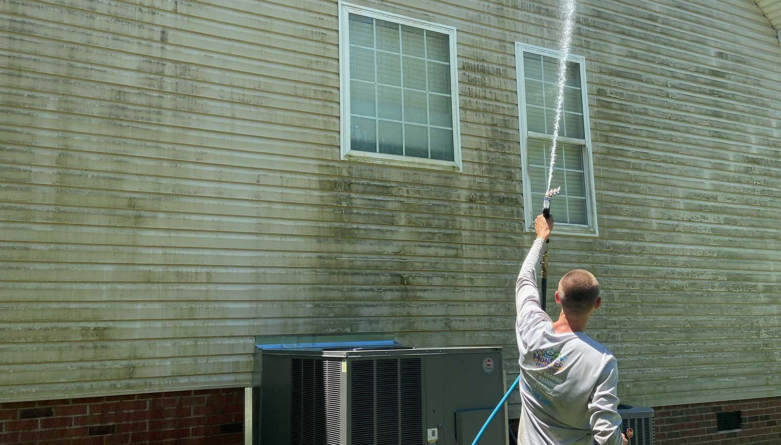 A person pressure washing the exterior side of a house, cleaning dirty, algae-covered siding near an air conditioning unit.