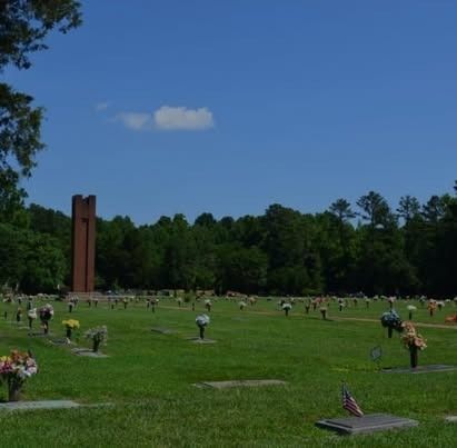 Lush green cemetery with headstones and bouquets. A large, brick-colored cross stands in the distance under a bright blue sky.