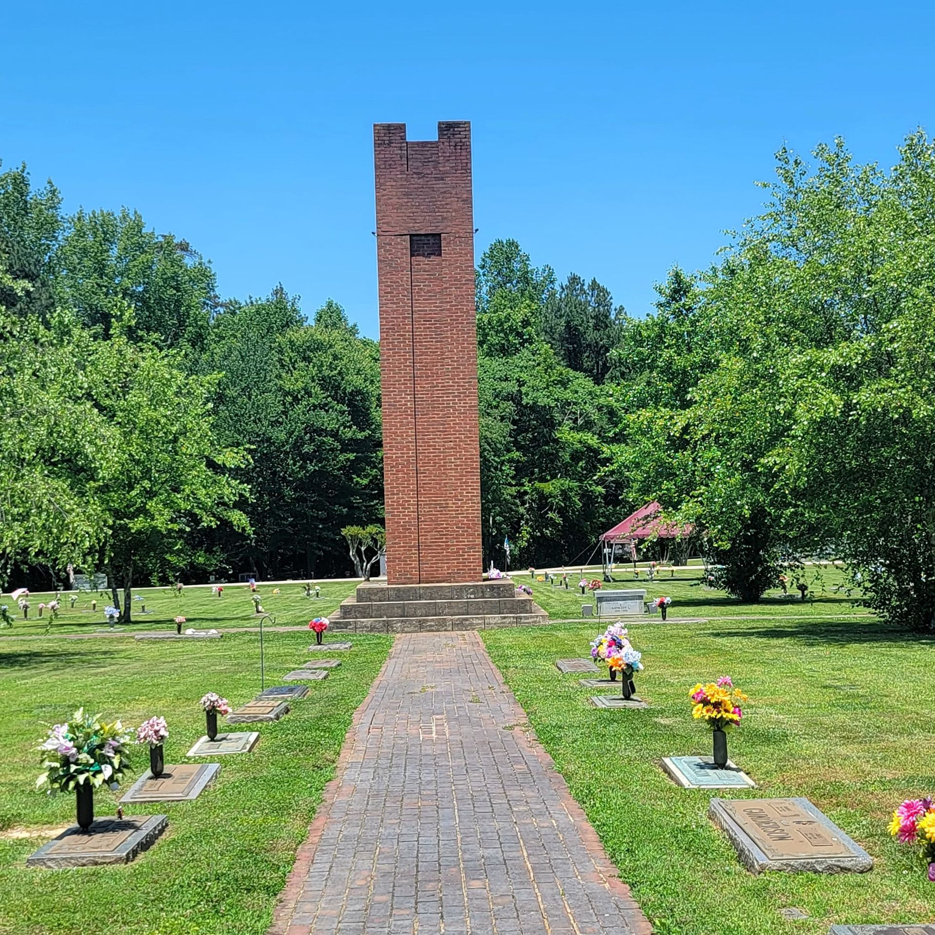 Brick tower monument in a cemetery with a brick path, tombstones, and trees under a blue sky.