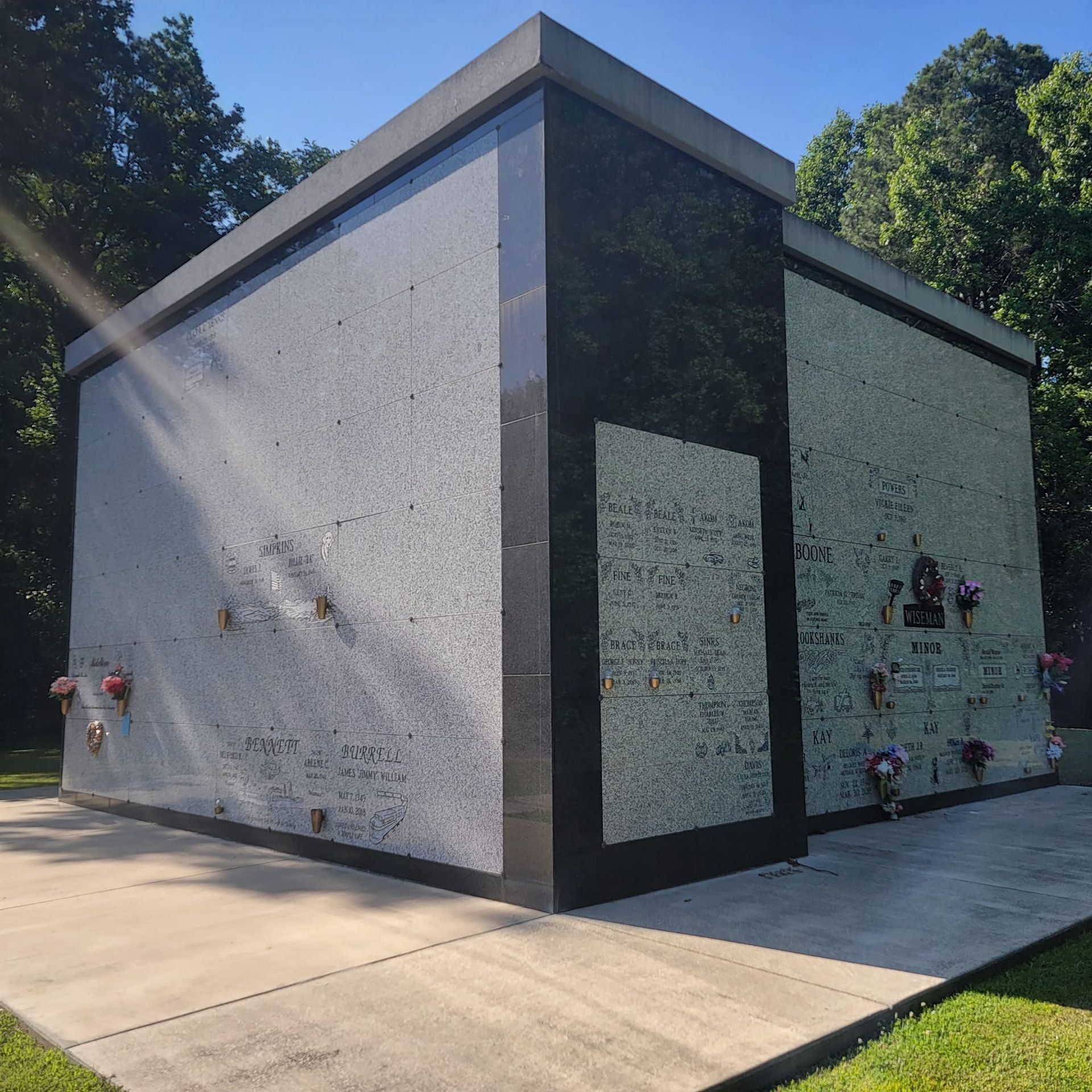 A mausoleum made of concrete with black trim and a concrete pathway in front. Flowers rest on the walls.