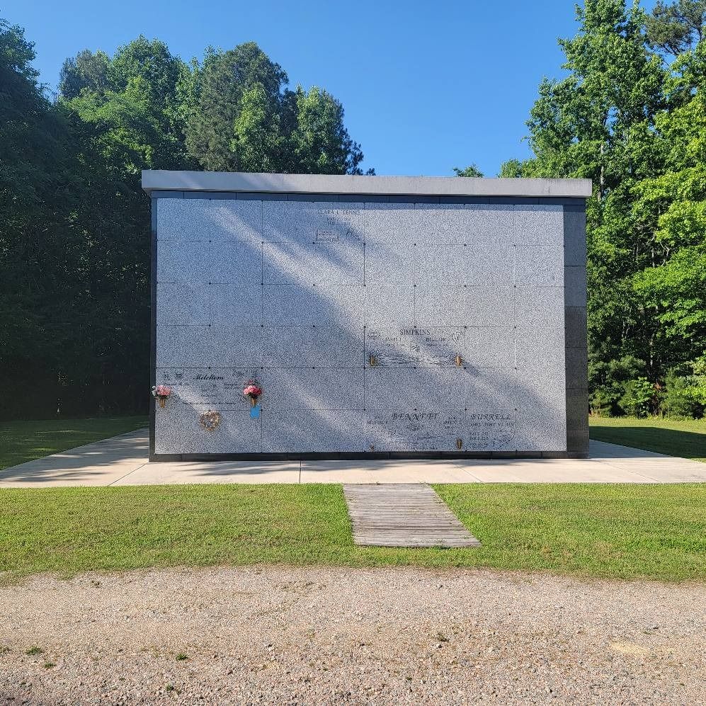 A granite columbarium in a cemetery with a grassy lawn, trees, and blue sky.