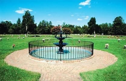 A cemetery with a fountain. Green grass, brick pathway, black fence, trees, and a blue sky.