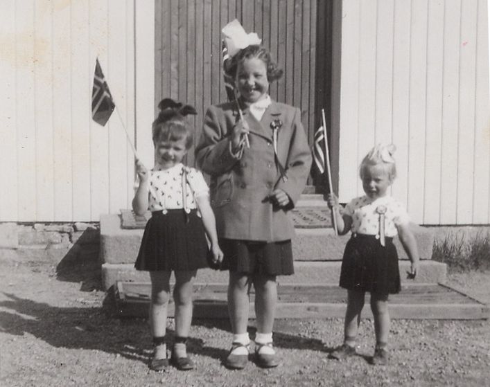 Tre barn i skjørt og bluser, som holder flagg og har på seg festhatter foran en trebygning.