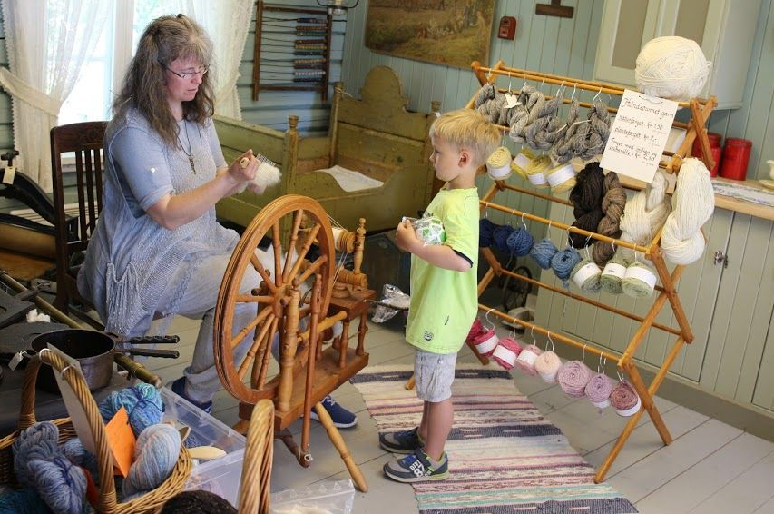 Kvinne spinner ull, barn ser på. Inne, gammelt hus, garn utstilt.