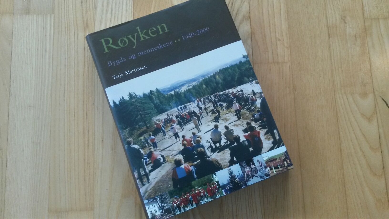Bok med tittelen «Røyken» på tregulv, med mennesker og natur på omslaget.