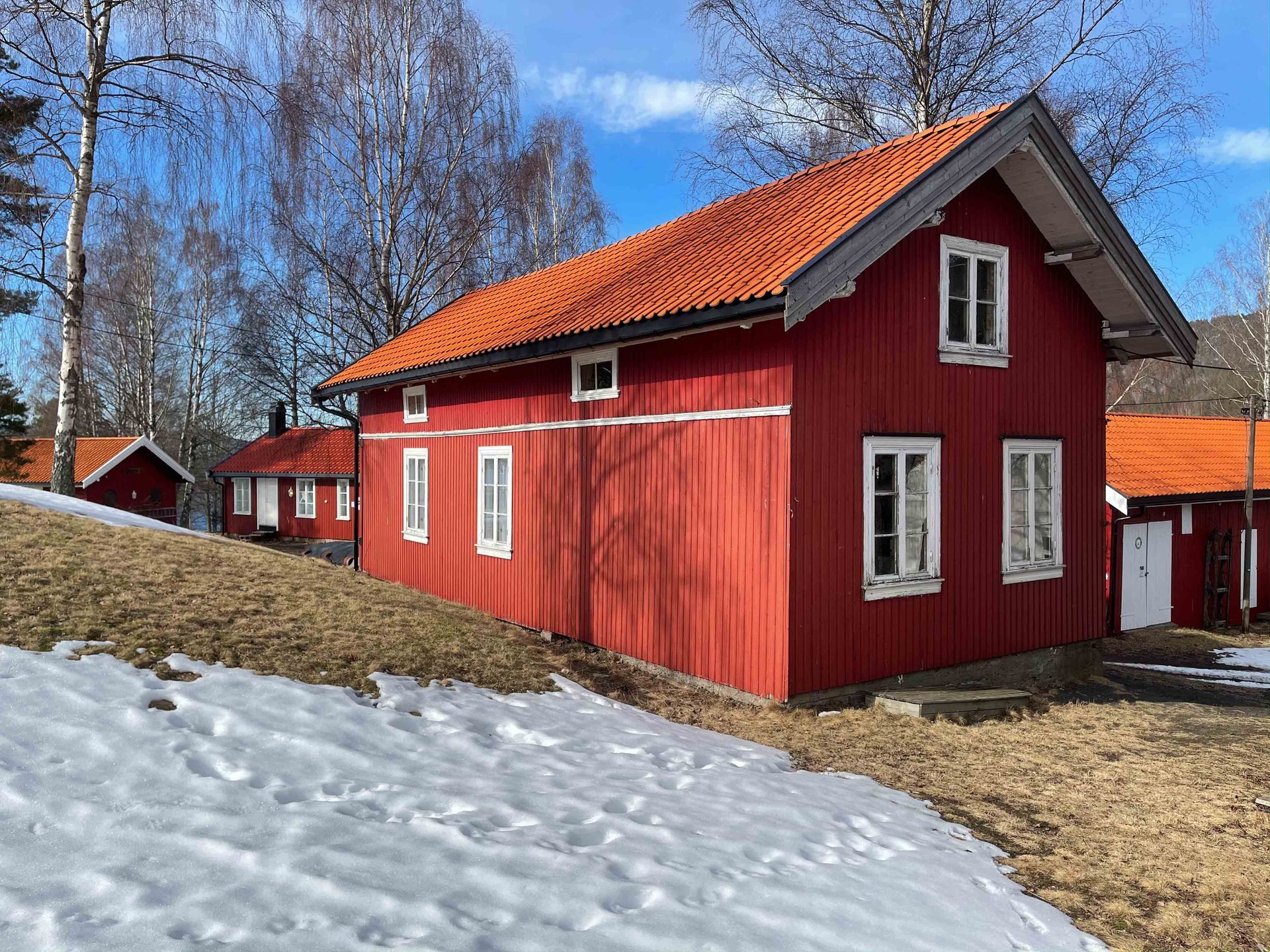 Røde trebygninger med hvite vinduer under en knallblå himmel, delvis dekket av snø.