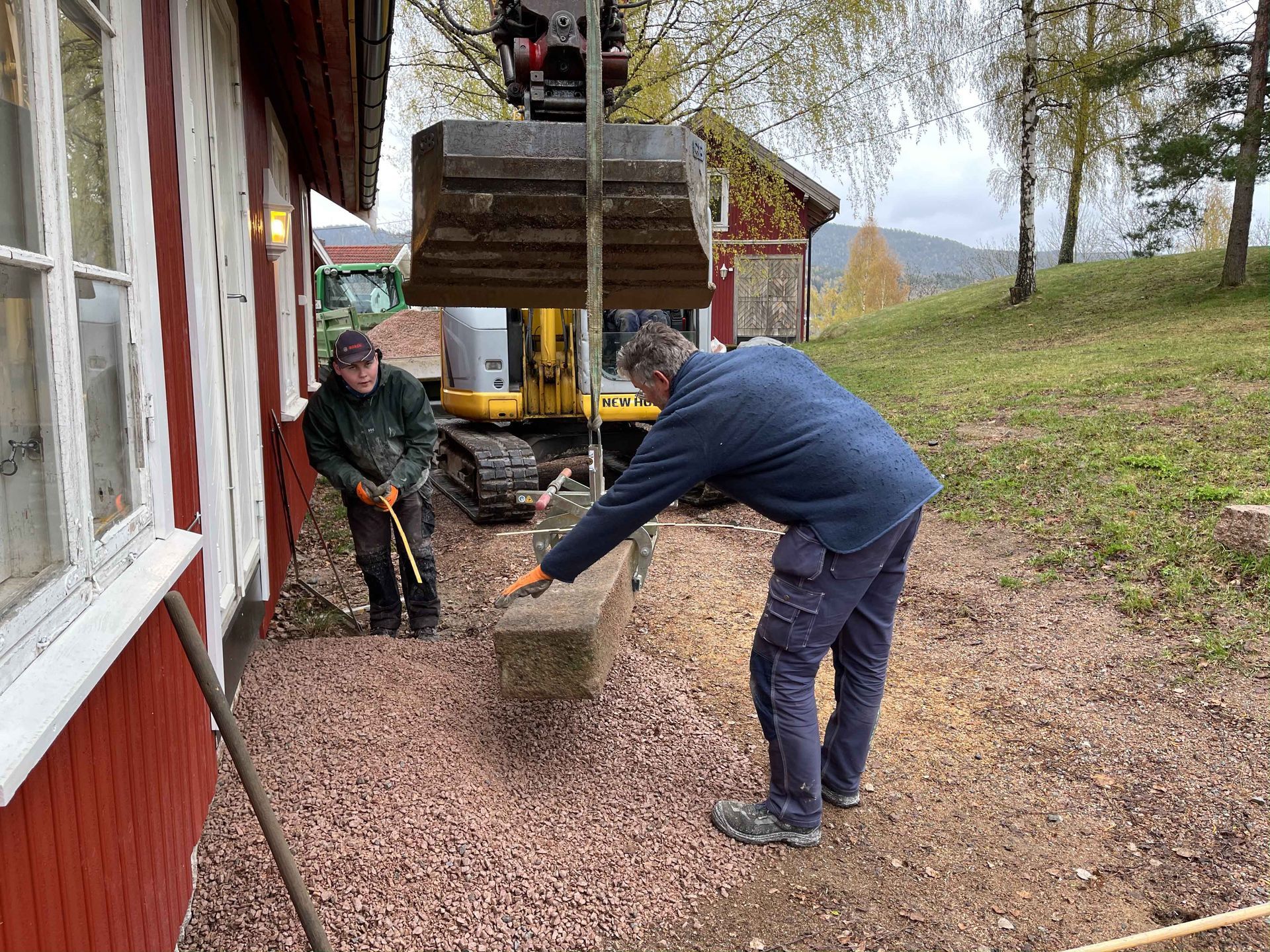 To menn og en gravemaskin jobber med grusfundamentet til et hus. Den ene mannen styrer grusen, den andre raker.