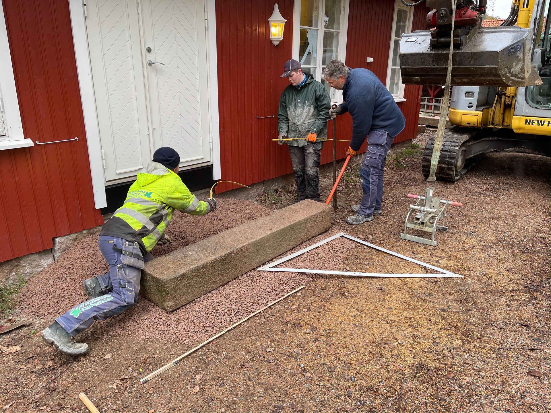 Arbeidere monterer en stor steinblokk ved siden av en rød bygning, assistert av en gravemaskin.